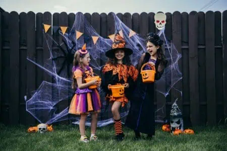Cheerful girls in costumes holding candy buckets standing in front of the fence with halloween decoration