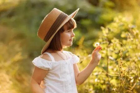 Adorable little girl wearing summer straw hat smelling wild flower at the park