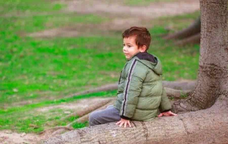 Young boy wearing jacket sitting on a tree log at the park