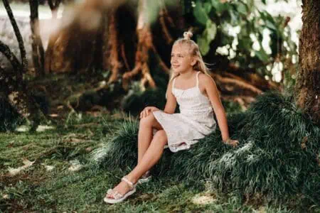 Beautiful young girl in white dress sitting on grassy tree roots in the forest