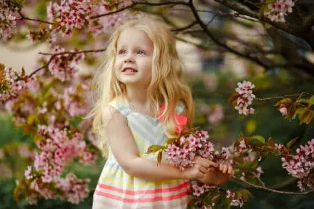 Blonde adorable girl wearing dress standing near spring tree with flowers