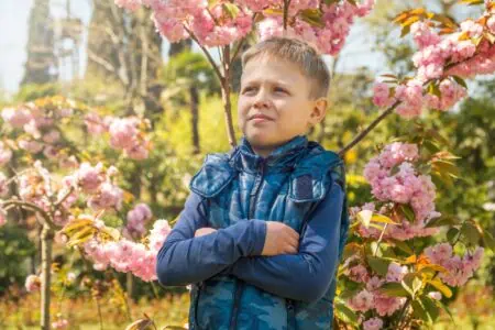 Handsome young boy wearing jacket posing with arms crossed in front of tree with blooming flowers at the park
