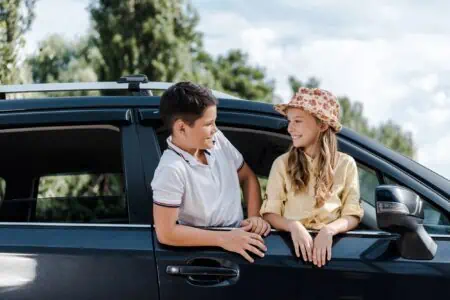 Adorable girl wearing a straw hat, looking at her brother while peeking through the car window, with only their upper half visible.