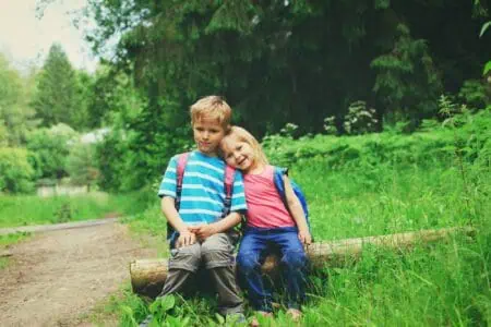 Cute siblings with backpacks sitting on a tree log in the meadow while resting